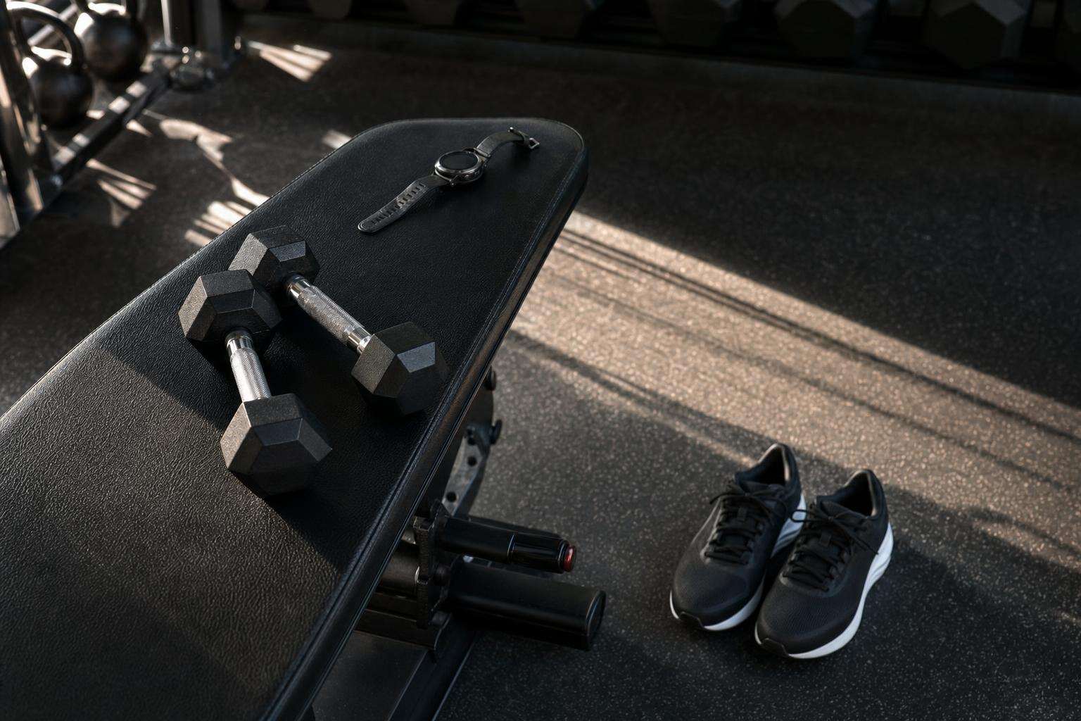 Dumbbells beside running shoes and a heart-rate watch on a bench, no people