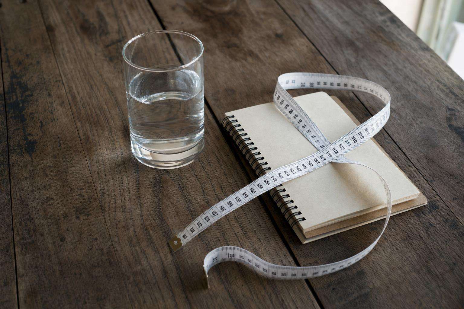 Measuring tape and notebook on a wooden table beside a glass of water, no people